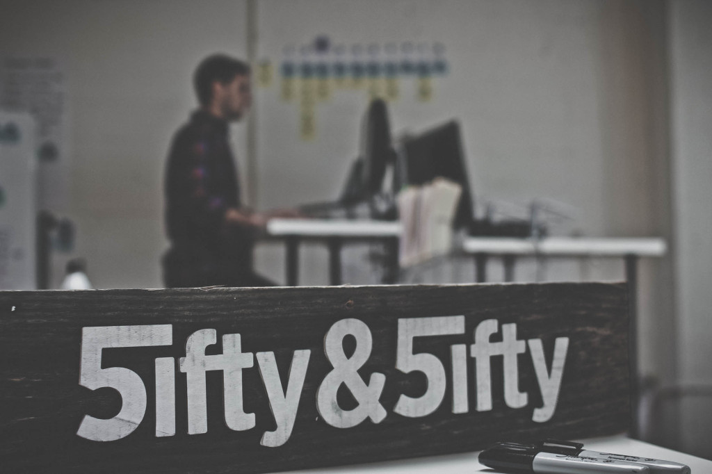 drewniana tablica ozdobiona napisem Fifty & Fifty zapisanym białymi literami jako „5ifty & 5ifty”. mężczyzna pracuje na komputerze w tle