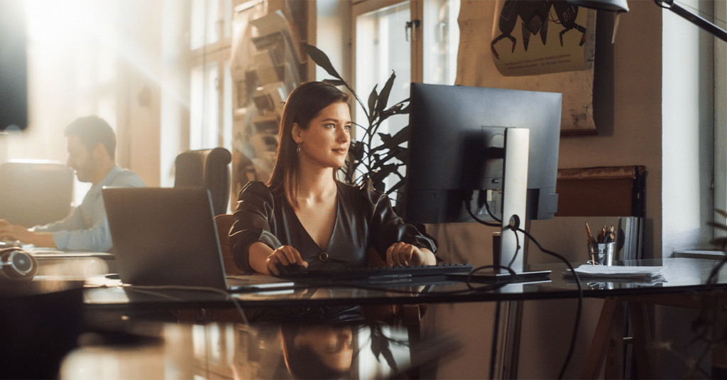 Frau arbeitet im Büro am Computer, Mann arbeitet im Hintergrund am Computer